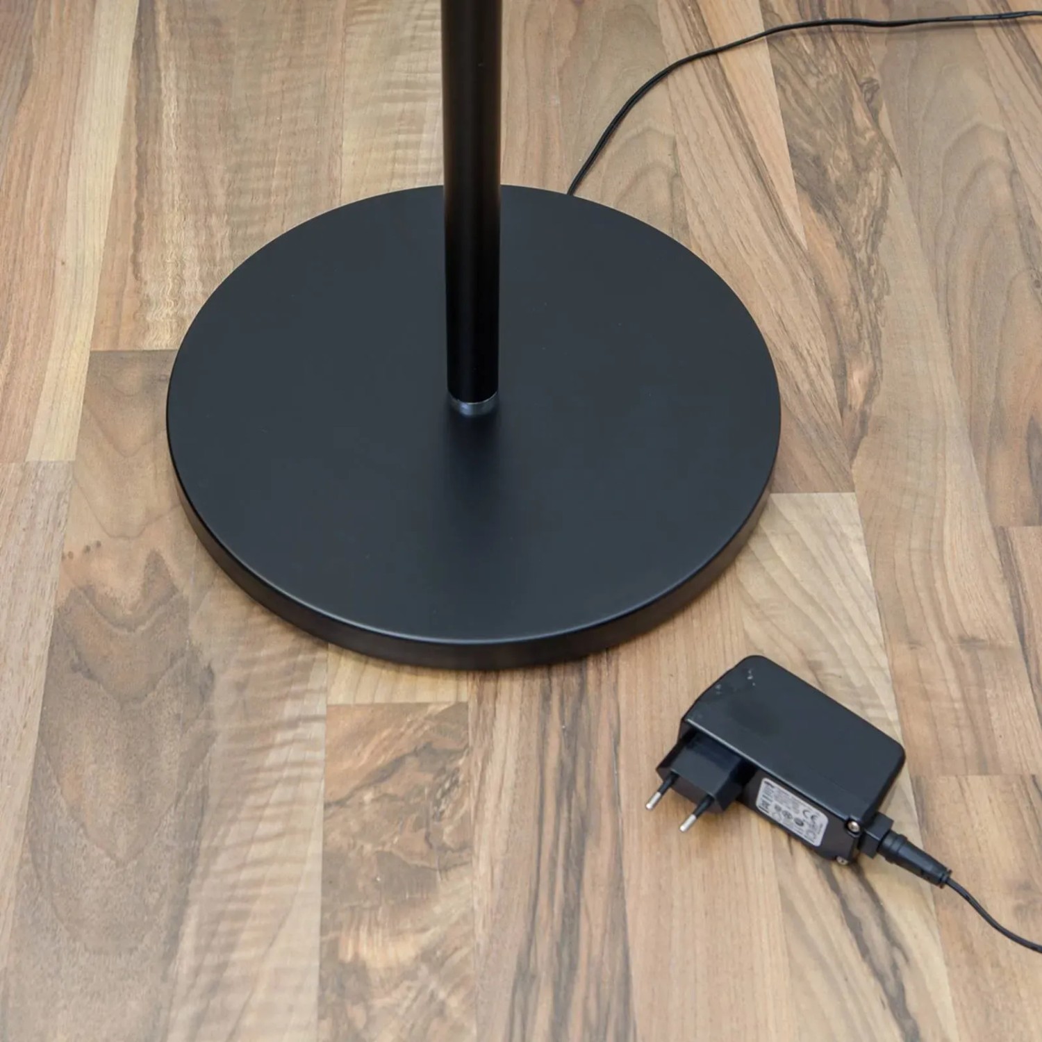 Detailaufnahme: Schwarze Lindby LED Stehlampe Malea mit rundem Fuß und Netzteil auf Holzboden.