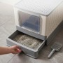 Weiße, geschlossene Katzentoilette aus Kunststoff mit grauer Schublade voller Streu, die herausgezogen wird. Graue Streuschaufel daneben.