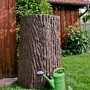 Arves Regentonne Evergreen 475 l in Dunkelbraun mit Baumstammoptik und grüner Gießkanne im Garten.
