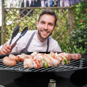 Schwarzer tragbarer Kugelgrill mit Grillrost voller Würstchen und Spieße; lächelnder Mann mit Grillzange im Garten.
