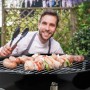 Schwarzer tragbarer Kugelgrill mit Grillrost voller Würstchen und Spieße; lächelnder Mann mit Grillzange im Garten.