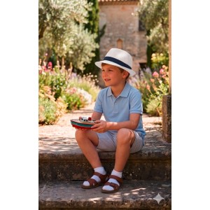 Junge mit weiß-blauem Fedora-Strohhut sitzt im Garten, hält Spielzeugboot. Kinder-Sonnenhut Gr. 53-54.