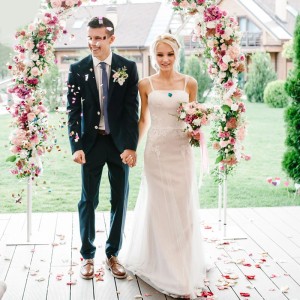 Glückliches Brautpaar unter Blumenbogen, umgeben von fallenden weißen Rosenblättern. Romantische Konfettikanone für Hochzeiten.