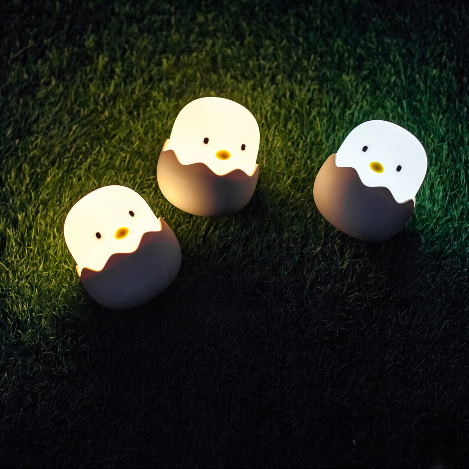 Drei dimmbare MegaLight LED Nachtlichter Eggy Egg in Kükenform auf Rasen.