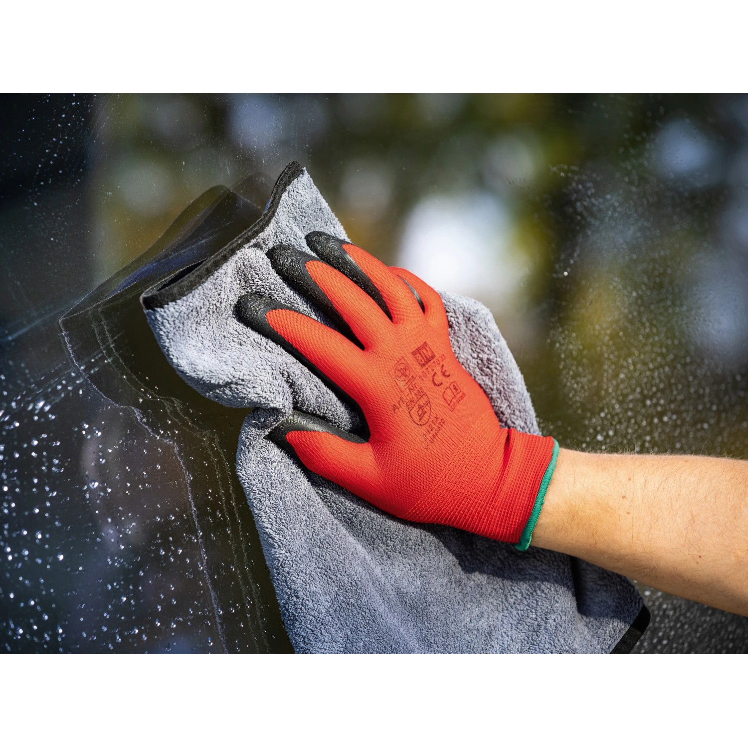 Hand mit rotem Arbeitshandschuh und Latex-Beschichtung reinigt eine Scheibe mit einem Tuch.