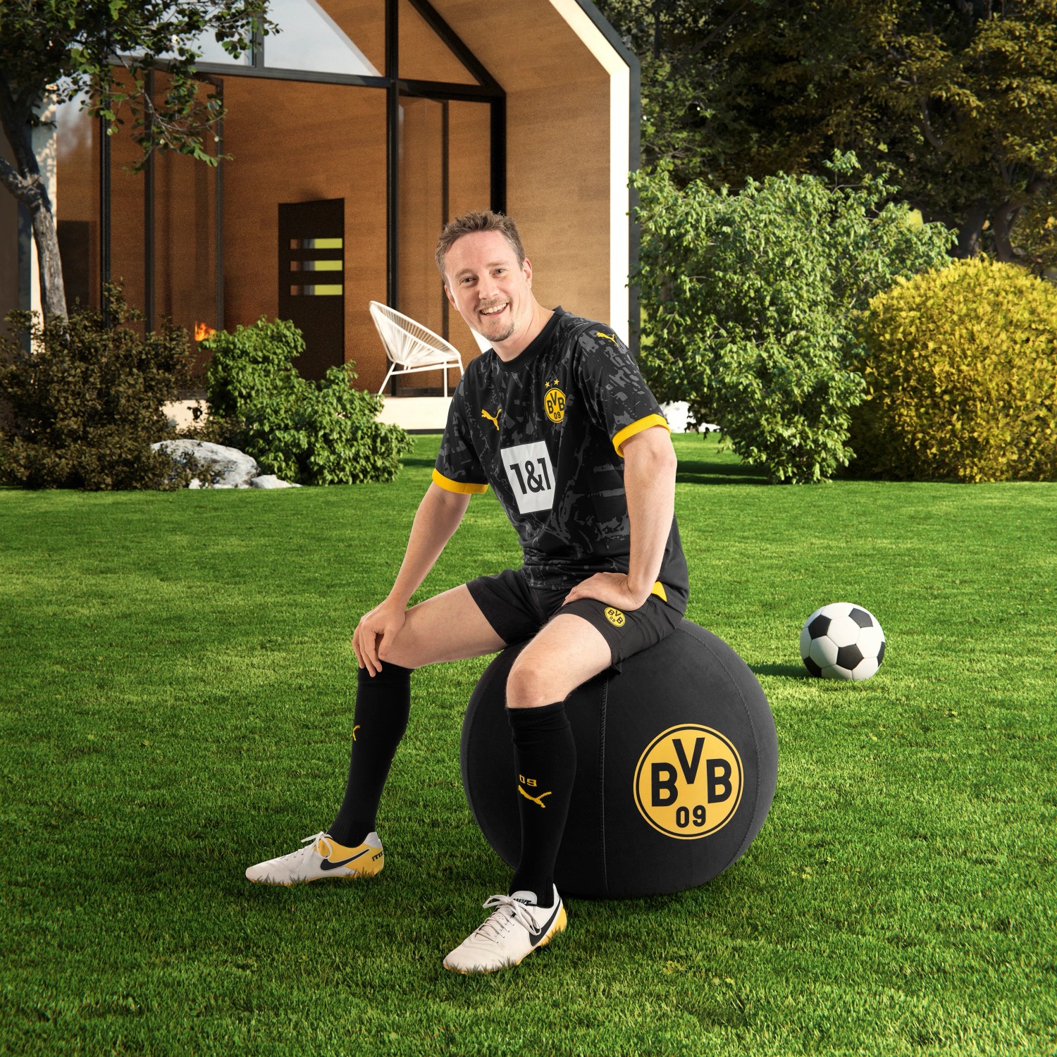 Mann sitzt auf schwarzem BVB Sitting Ball Hocker 120l im Garten.