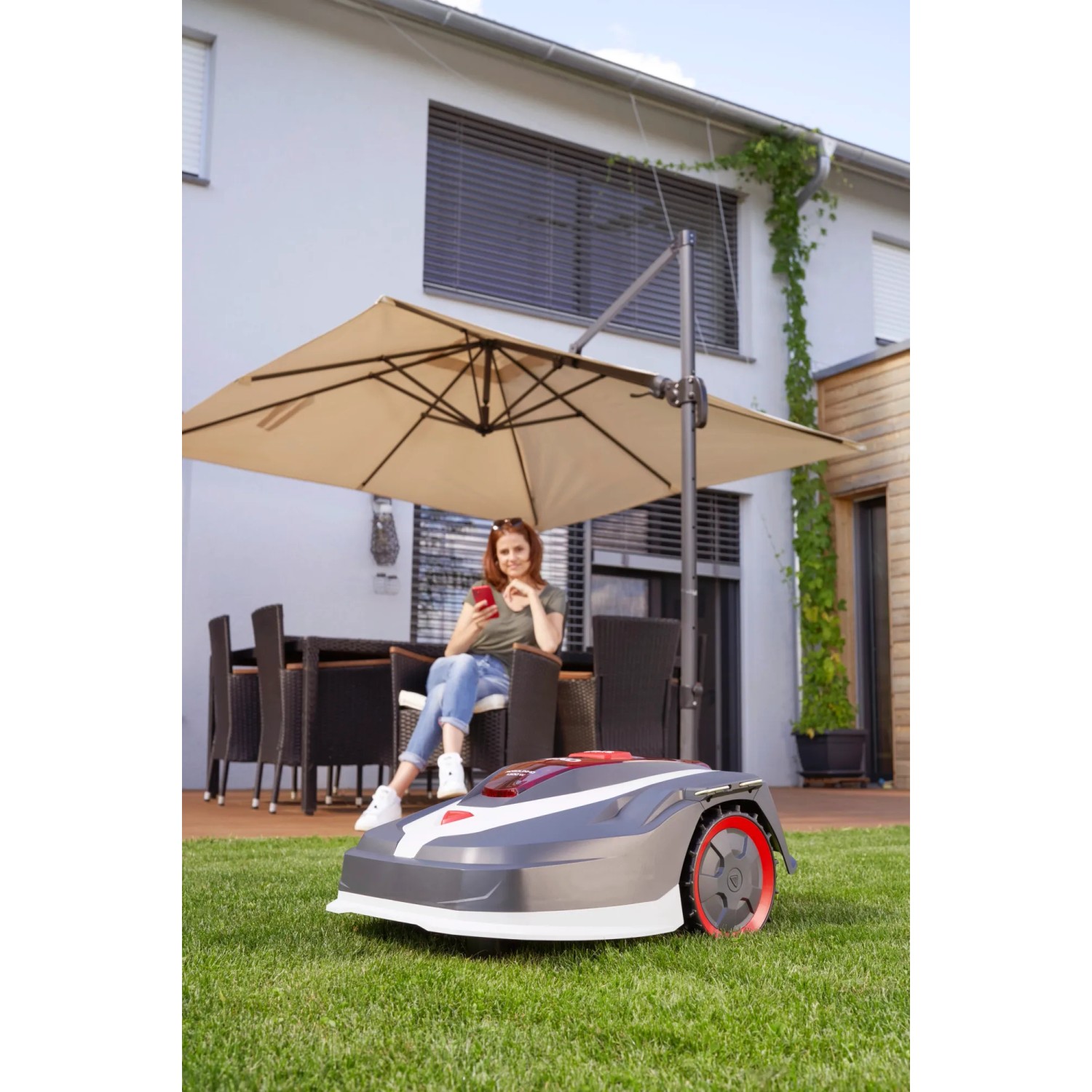 Al-Ko Robolinho 1.300 W Mähroboter im Garten mit Frau unter Sonnenschirm.