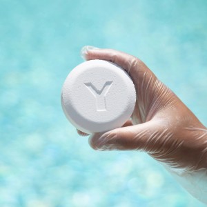 Hand mit Schutzhandschuh hält Bayrol Chlorilong Power 5 Chlortablette vor Pool.