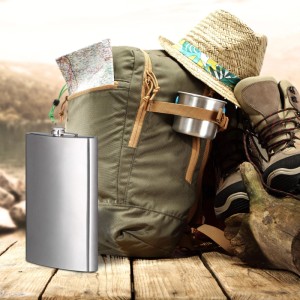 Großer silberner Edelstahl-Flachmann (1,8L) für Camping & Outdoor, neben Rucksack und Wanderschuhen auf Holz.