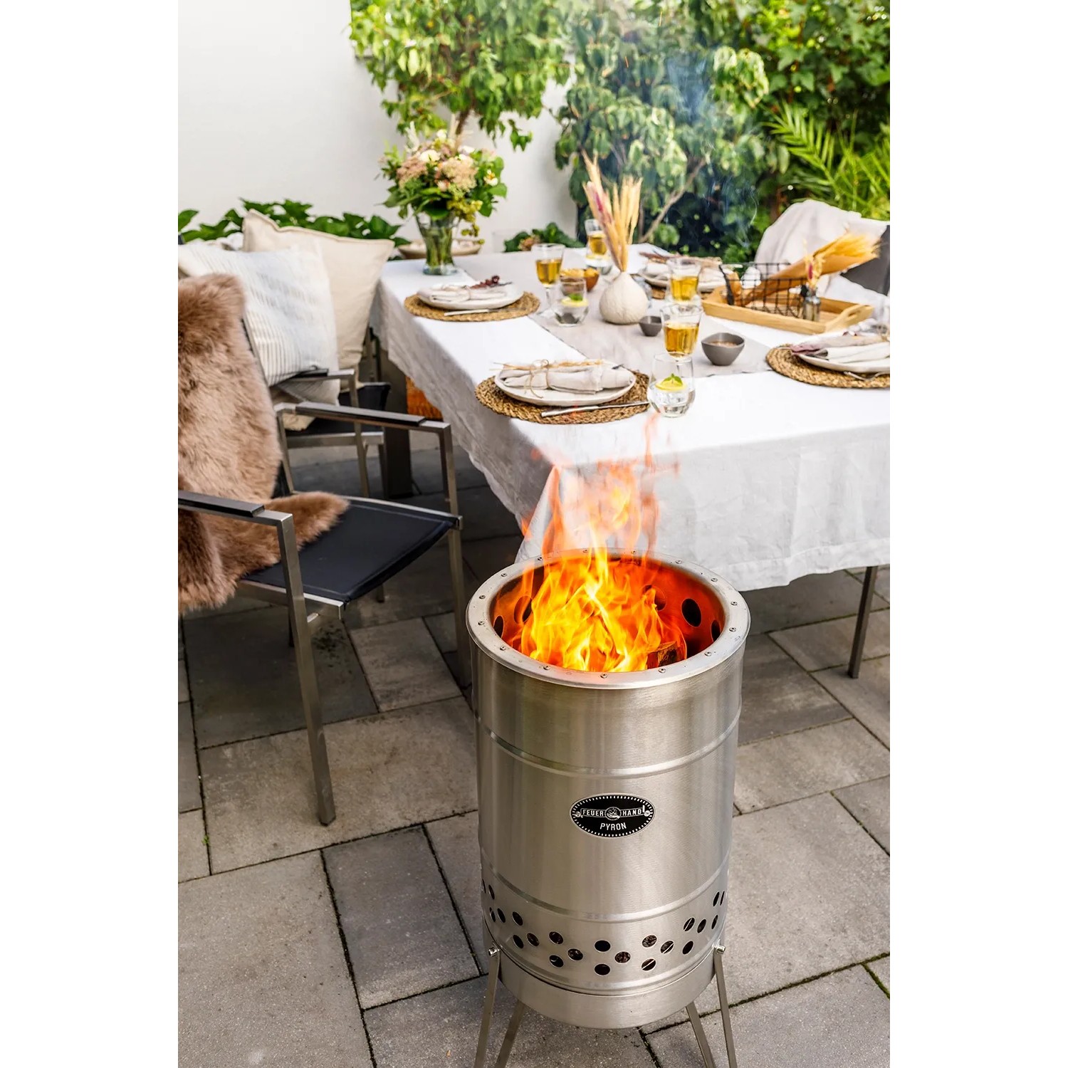 Feuerhand Pyron Feuertonne aus Edelstahl, 25 l, mit brennendem Holz vor gedecktem Tisch im Garten.