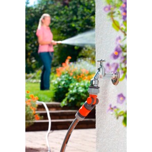 Gardena Winkelhahnstück am Wasserhahn mit Schlauch verbunden. Frau gießt im Hintergrund den Garten.