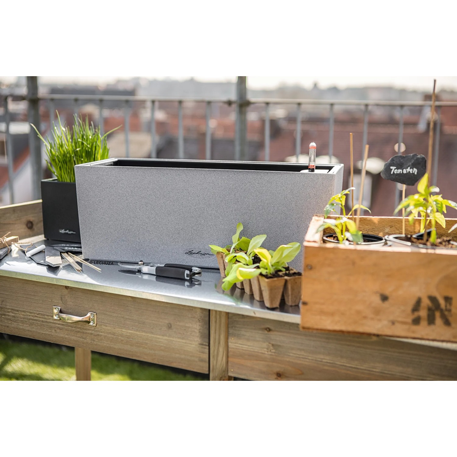 Lechuza Balconera Stone 50 Blumenkasten, steingrau, mit Pflanzen und Zubehör auf einem Gartentisch.