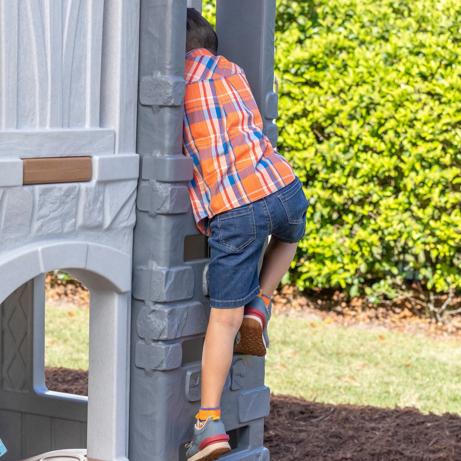 Kind klettert auf ein graues Step2 Spielhaus Adventures mit Rutsche im Garten.