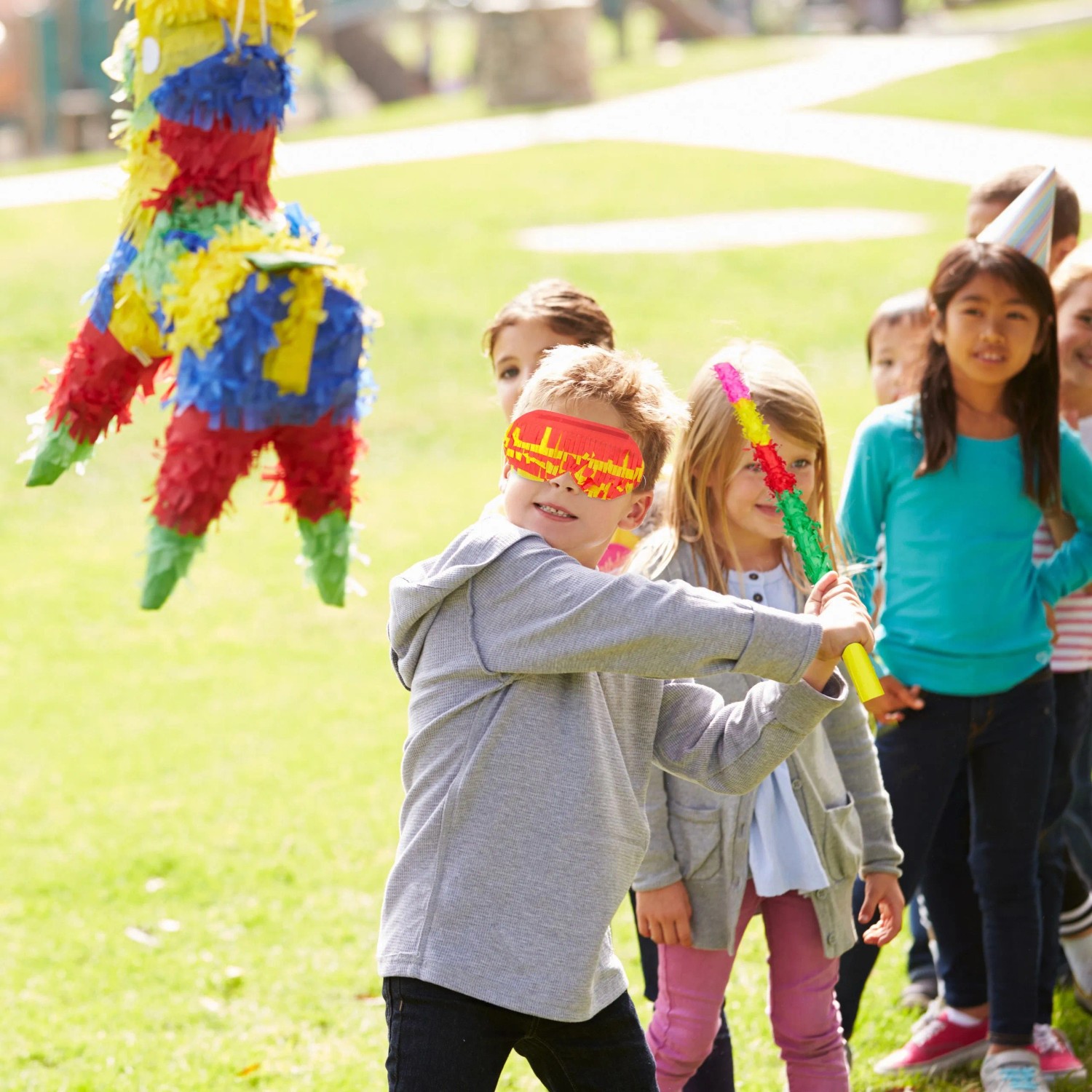 Kind mit Augenmaske schlägt mit buntem Pinata Stab auf Pinata. Pinata Zubehör Set für Partys und Geburtstag.