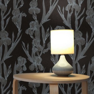 Schwarze Tadessi Vliestapete mit hellem floralem Iris-Muster, beleuchtet von einer Tischlampe auf Holztisch. Dekorative Tapete.