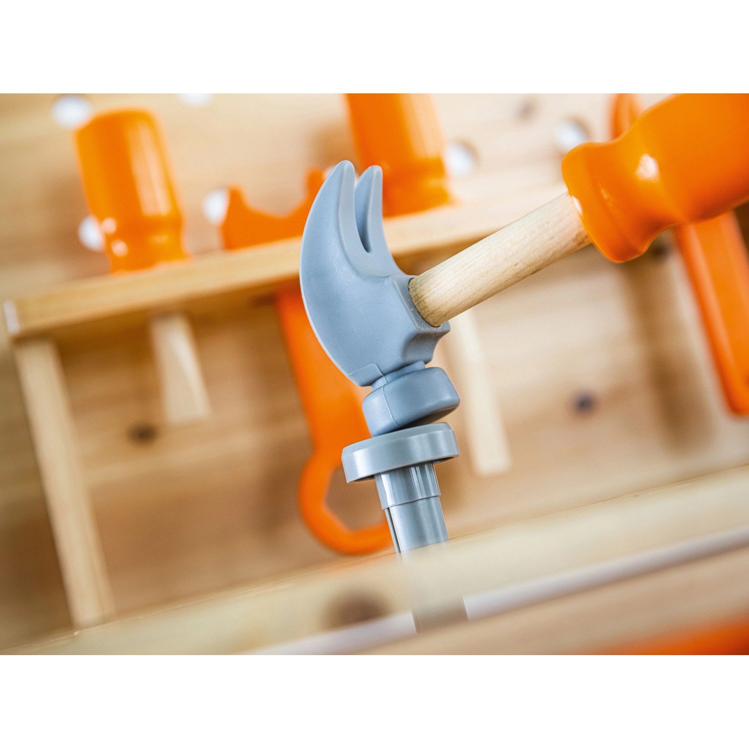 Kinderhammer schlägt grauen Steckstift in OBI Kinderwerkbank aus Holz. Orange Spielzeugwerkzeuge im Hintergrund.