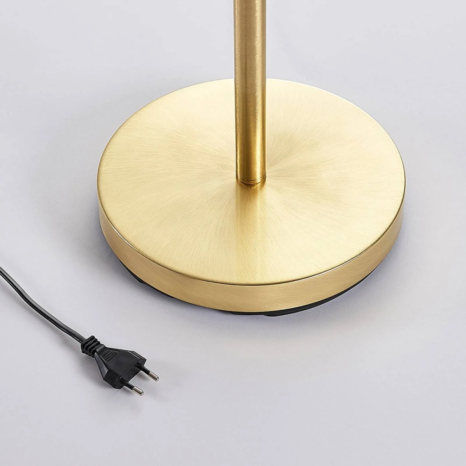 Runder goldener Messing Metallfuß der Lindby Stehlampe Jost. Schwarzes Stromkabel mit Stecker auf hellem Boden.