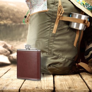 Brauner Edelstahl-Flachmann mit Kunstlederhülle und Bügelverschluss auf Holztisch vor Rucksack. Camping Zubehör.