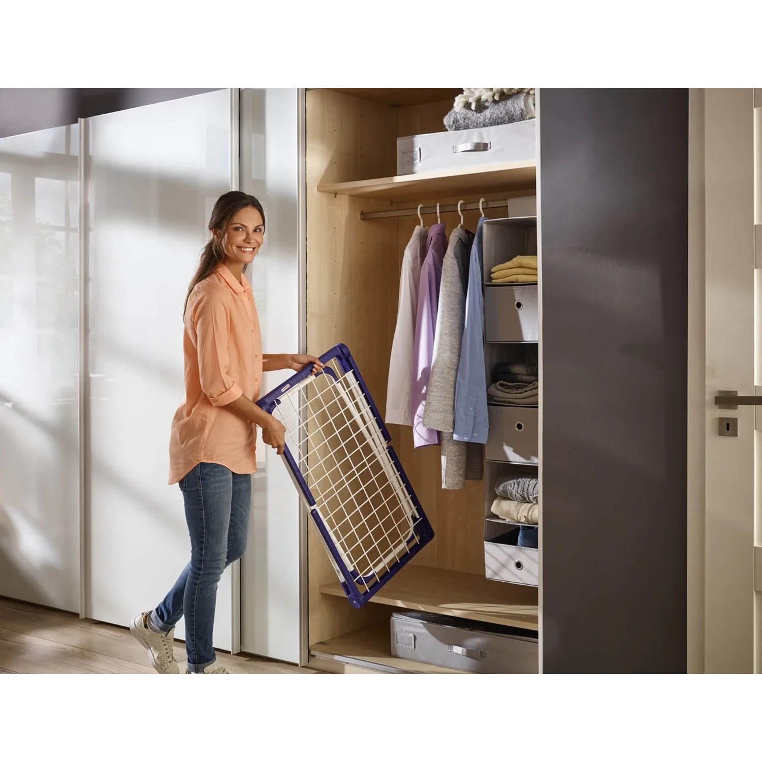 Frau hält zusammengeklappten Leifheit Wäscheständer Pegasus 120 Compact vor Kleiderschrank.