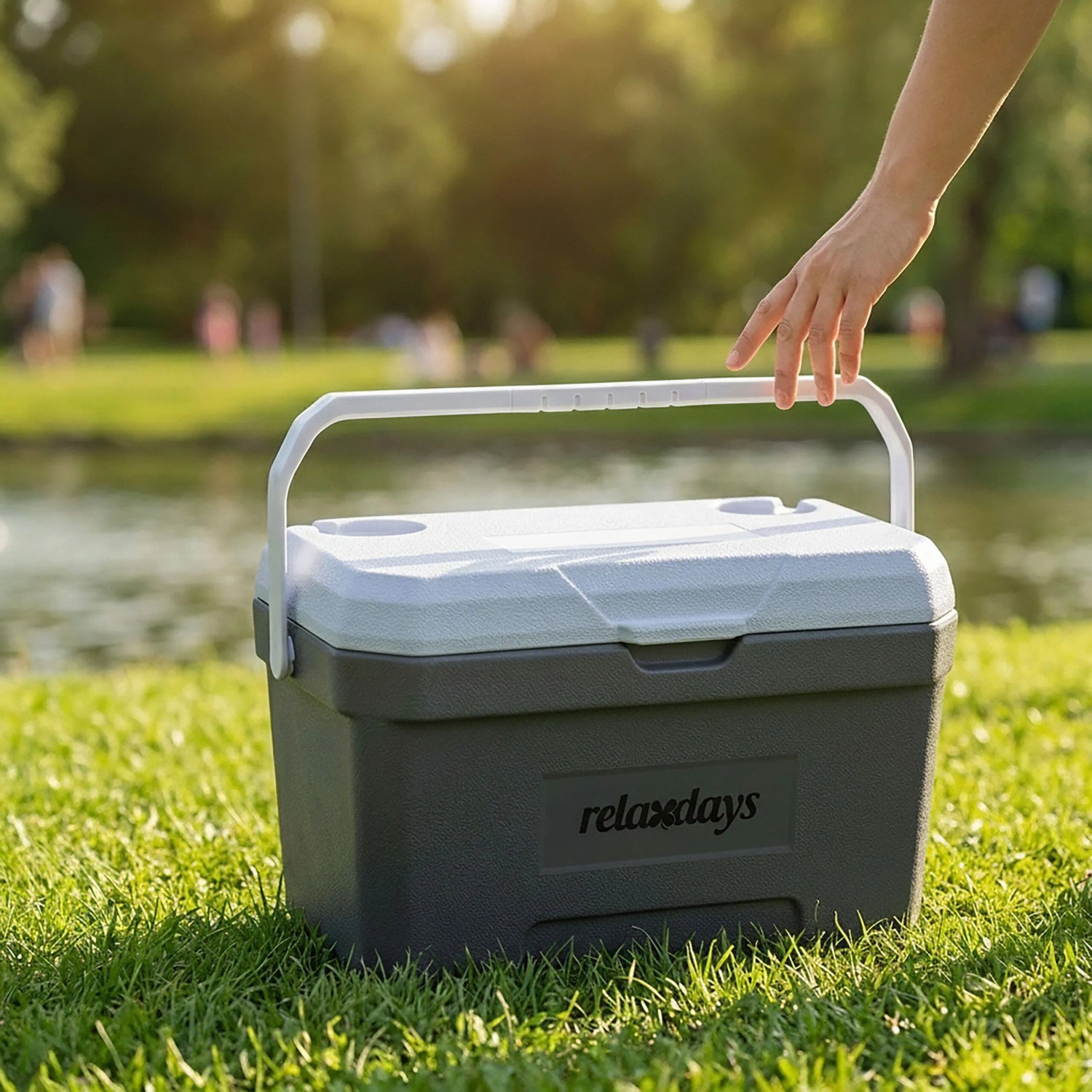 Graue Relaxdays Kühlbox aus Kunststoff mit weißem Deckel und Tragegriff auf grünem Gras am See. Für Picknick & Camping.