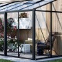 Modernes Bloomcabin Wandgewächshaus mit schwarzem Aluminiumrahmen, Glaswänden, blühenden Pflanzen und Sitzbereich an Holzwand.