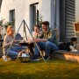 Petromax Dreibein mit Kette und Haken über Feuerstelle mit Familie beim Grillen im Garten.