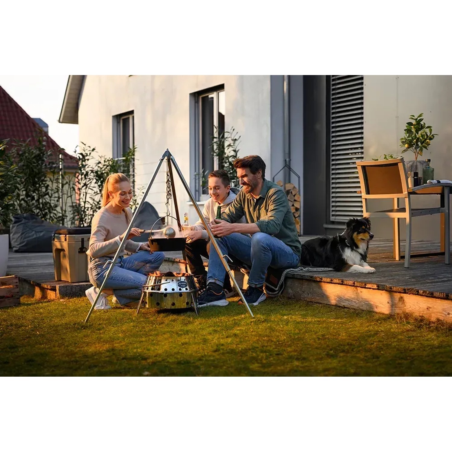 Petromax Dreibein mit Kette und Haken über Feuerstelle mit Familie beim Grillen im Garten.