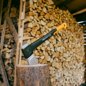Fiskars Spaltaxt X24 X-series M auf Holzscheit vor Holzstapel. Axt mit schwarzem Stiel und orangefarbenem Griff.