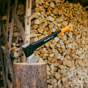 Fiskars Spaltaxt X24 auf Holzscheit vor Holzstapel. Axt mit schwarzem Stiel und orangefarbenem Griff.