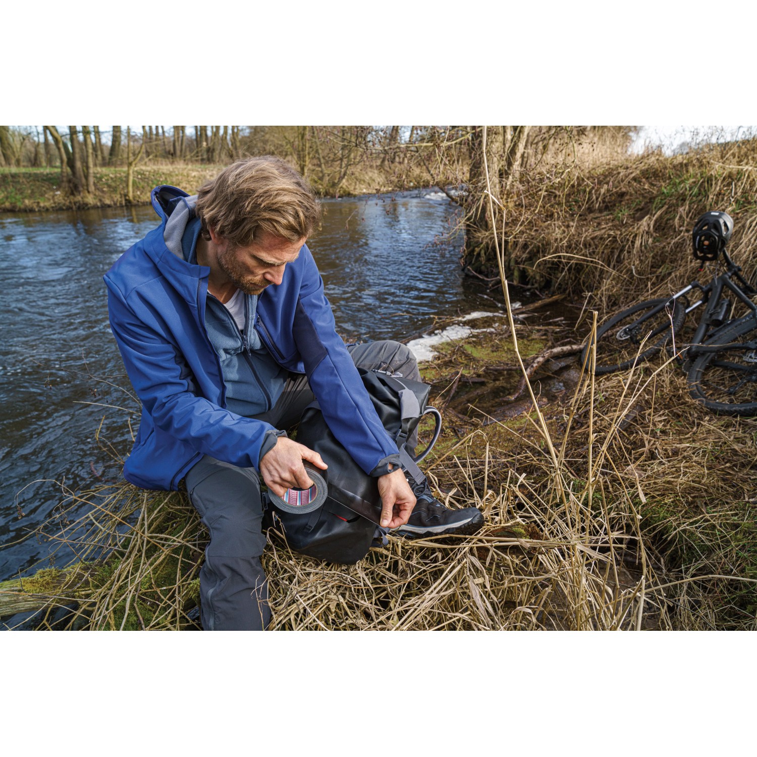 Mann repariert Rucksack mit Tesa Ultra Power Extreme Reparaturband am Fluss.