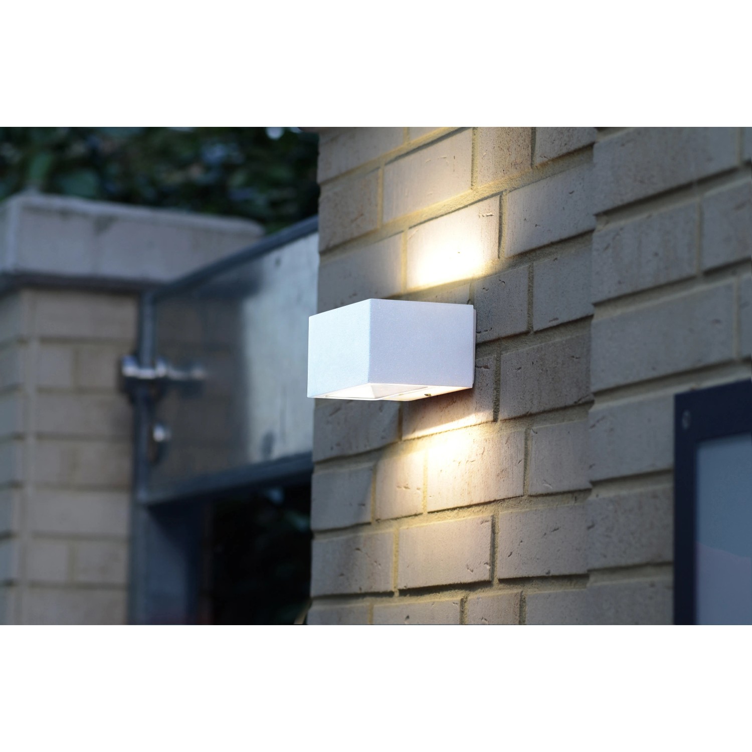 Weiße LUTEC LED-Außenwandleuchte Gemini an einer Backsteinmauer, die Licht nach oben und unten abstrahlt.