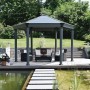 Anthrazitfarbener Palram Canopia Gartenpavillon Roma 4.1 x 3.6 m mit Sitzgruppe am Teich.