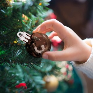Glänzende braune Weihnachtskugel aus Kunststoff wird von Kinderhand an grünen Weihnachtsbaum gehängt. Festliche Christbaumdeko.