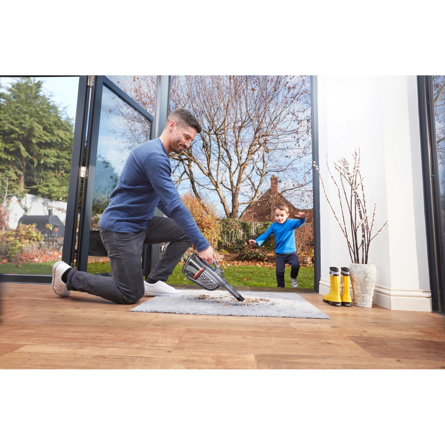 Mann saugt mit Black+Decker Akku-Handstaubsauger Teppich; Kind im Hintergrund.