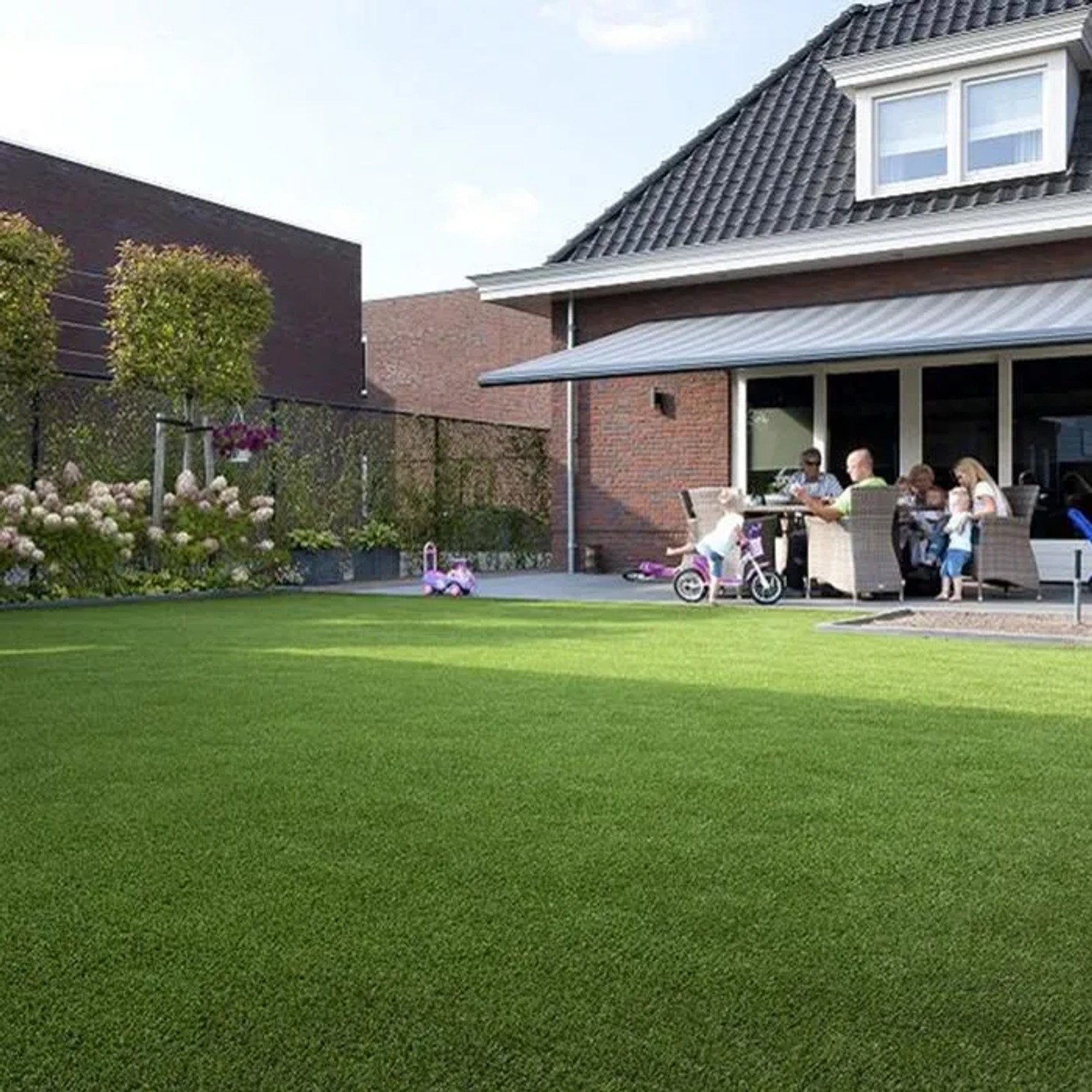 Sonniger Garten mit Karat Kunstrasen Nantes, 40mm hoch, UV-beständig, mit spielenden Kindern.