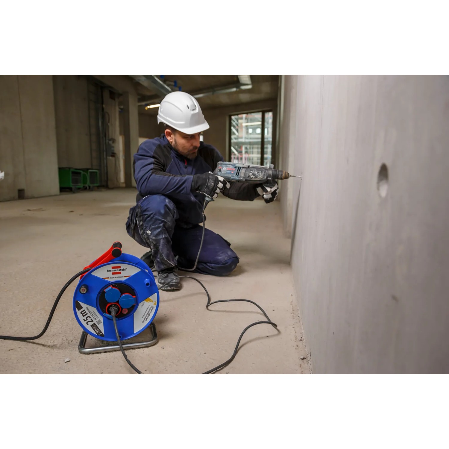 Blaue Garant Kabeltrommel 25m IP44 auf Baustelle, Arbeiter bohrt in Wand. Ideal für Gewerbe & Outdoor.