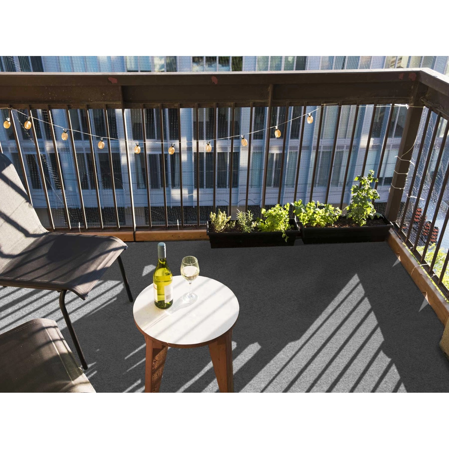 Grauer Primaflor Rasenteppich auf Balkon mit Tisch, Wein und Pflanzen. Kunstrasen für Terrasse.