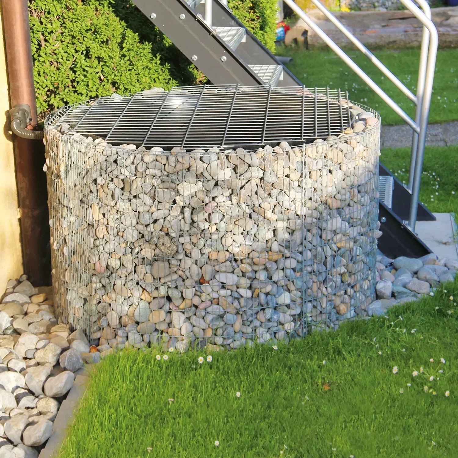 Gabionen-Wasserfass (750 l) im Garten, befüllt mit Steinen. Regentonne mit Gitterabdeckung.