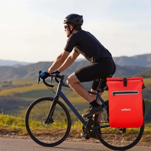 Rote Wiltec 20L Gepäckträgertasche am Fahrrad eines Radfahrers auf einer Straße in bergiger Landschaft.