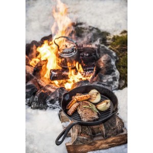 Petromax Grillpfanne aus Gusseisen mit Steak, Gemüse und Lagerfeuer im Hintergrund.