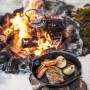 Petromax Grillpfanne aus Gusseisen mit Steak, Gemüse und Lagerfeuer im Hintergrund.