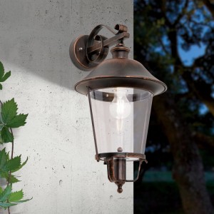 Rustikale Außenwandleuchte in Schwarz-Kupfer mit Laternen-Design und klarem Glas, leuchtend an einer Hauswand im Dunkeln.