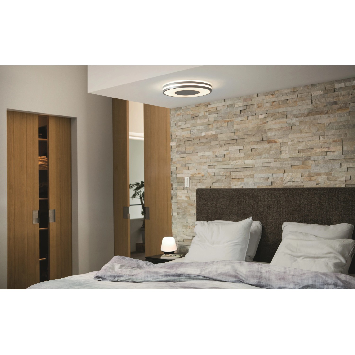 Silberne Philips Hue Being Deckenleuchte mit Dimmer im Schlafzimmer mit Steinwand und Holztüren.