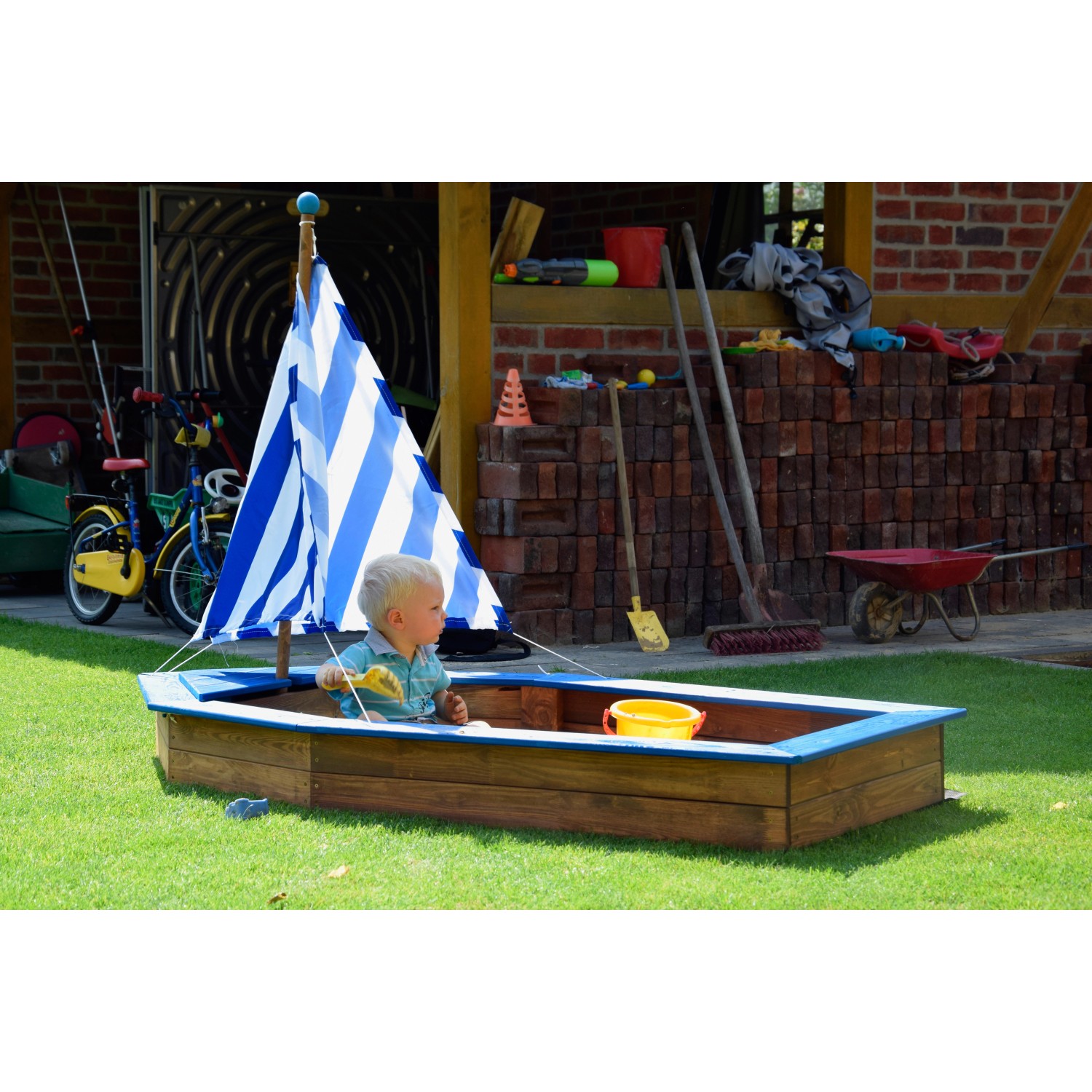 Dunkelbrauner Sandkasten im Schiff-Design mit blau-weißem Segel und spielendem Kind.