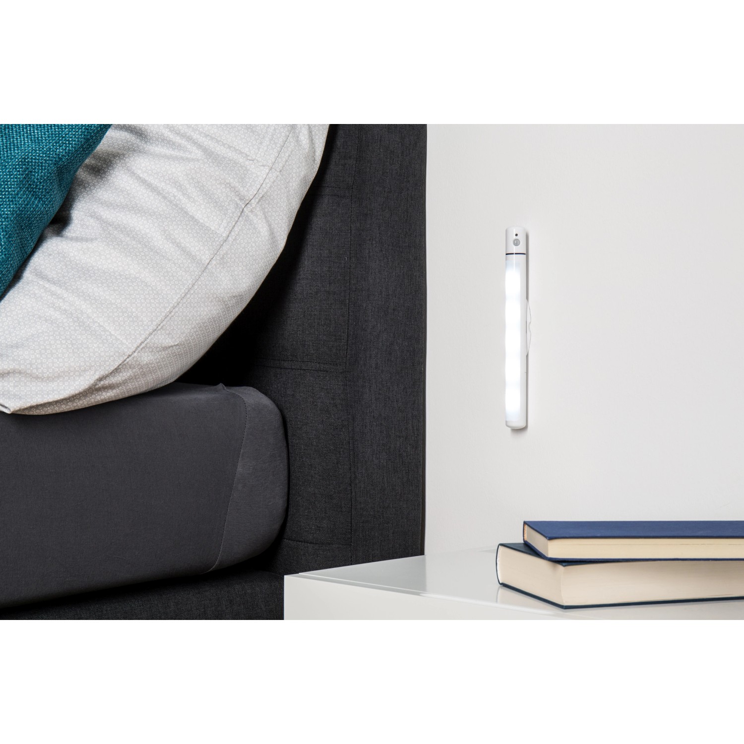 Weiße Müller-Licht LED Unterbauleuchte Falis mit Sensor an einer Wand über einem Nachttisch.