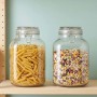 Zwei transparente Einmachgläser mit Bügelverschluss, gefüllt mit Nudeln und Müsli, auf Holzregal. Ideal für Küchenaufbewahrung.