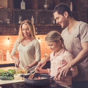 Lachende Familie kocht auf schwarzem Klarstein Induktionskochfeld. Vater, Tochter rühren in Pfanne, Mutter schnippelt Gemüse.