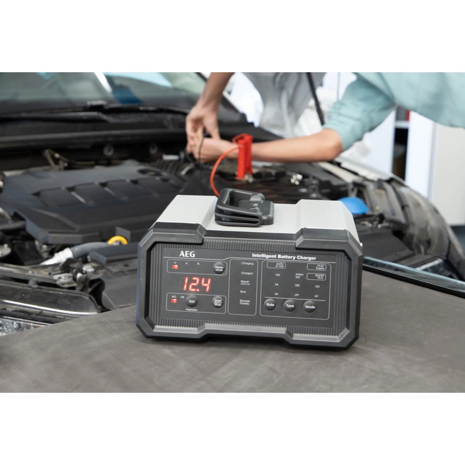 AEG Batterieladegerät CW10 vor einem Auto mit angeschlossenen Batterieklemmen.