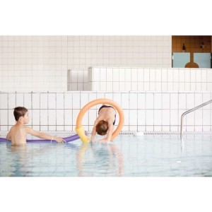 Jungen spielen im Pool mit gelber NMC Poolnudel. Ein Kind taucht durch die Schwimmhilfe.