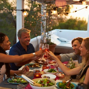 Fröhliche Menschen stoßen im Garten mit Getränken an, im Hintergrund Gartenstuhlauflagen.